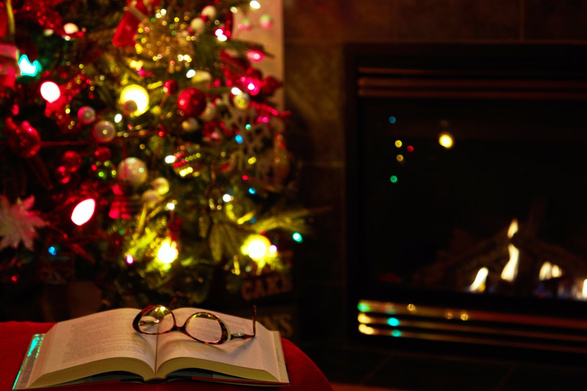 photograph of an open book with a pair of reading glasses resting on the pages. They are on a table behind which we can see a lit up Christmas tree and a the flickers of a fire next to it.  