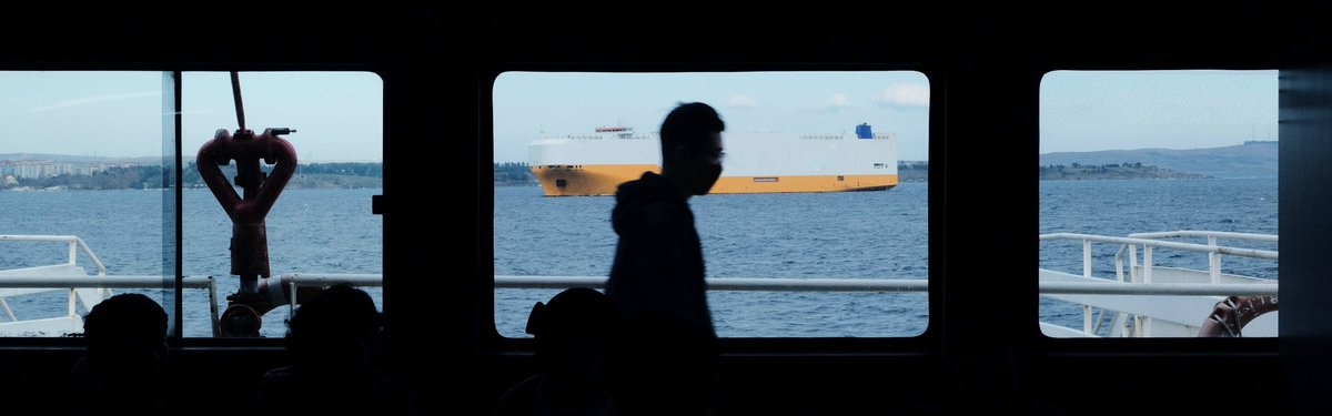 Darkened view of a masked seafarer standing on a ship in front of a window. There is a yellow and white car carrier type of ship on the water visible from the window. 