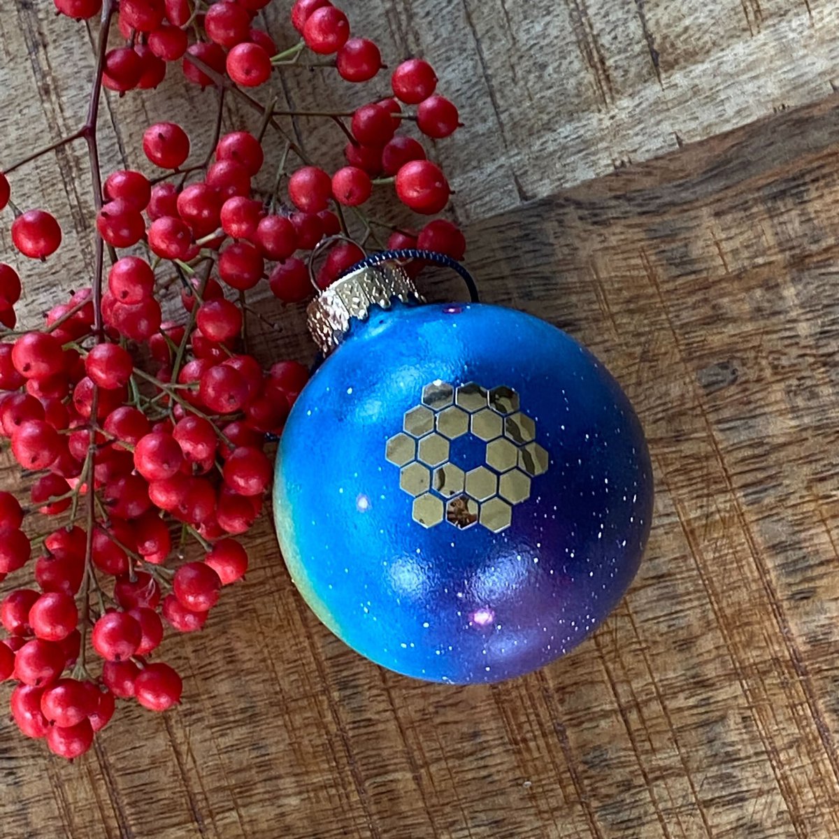 Nebula-painted glass globe ornament with the JWST gold hexagonal segmented mirror, on a wood background with red berries in the corner of the frame