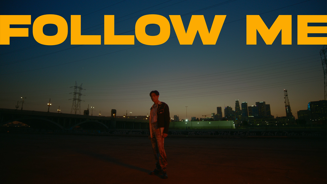 A nighttime scene shows a person standing in an empty lot with the city skyline behind them, below large yellow text reading 'FOLLOW ME'.