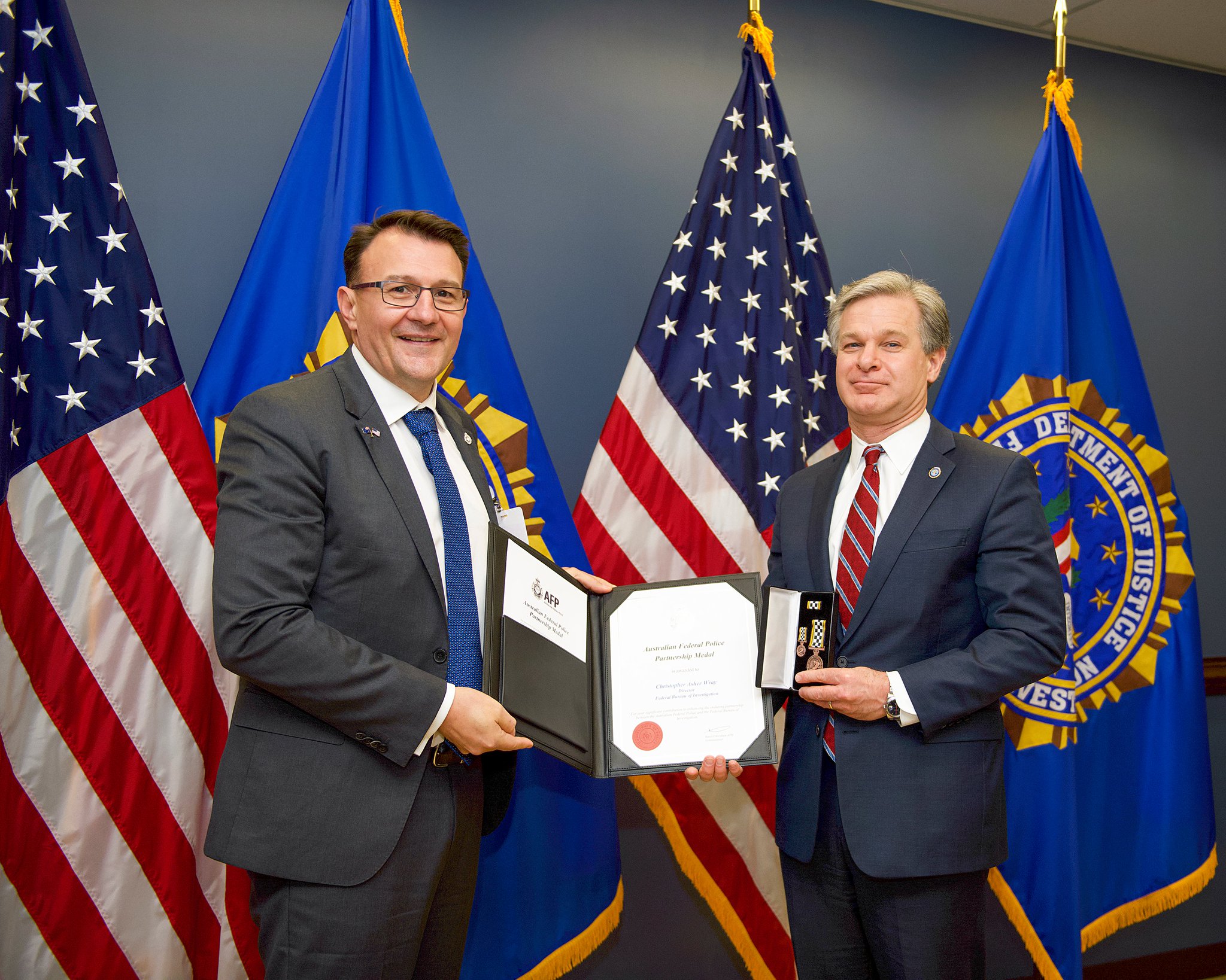 Australian Federal Police Commissioner Reece Kershaw (left) poses with Federal Bureau of Investigation Director Christopher Wray (right). Director Wray is holding a box containing large and small versions of the Australian Federal Police Partnership Medal in his left hand, and both men are holding a black folio with a corresponding certificate and paperwork. They are standing in front of American flags and FBI flags, which are displayed against a gray wall.