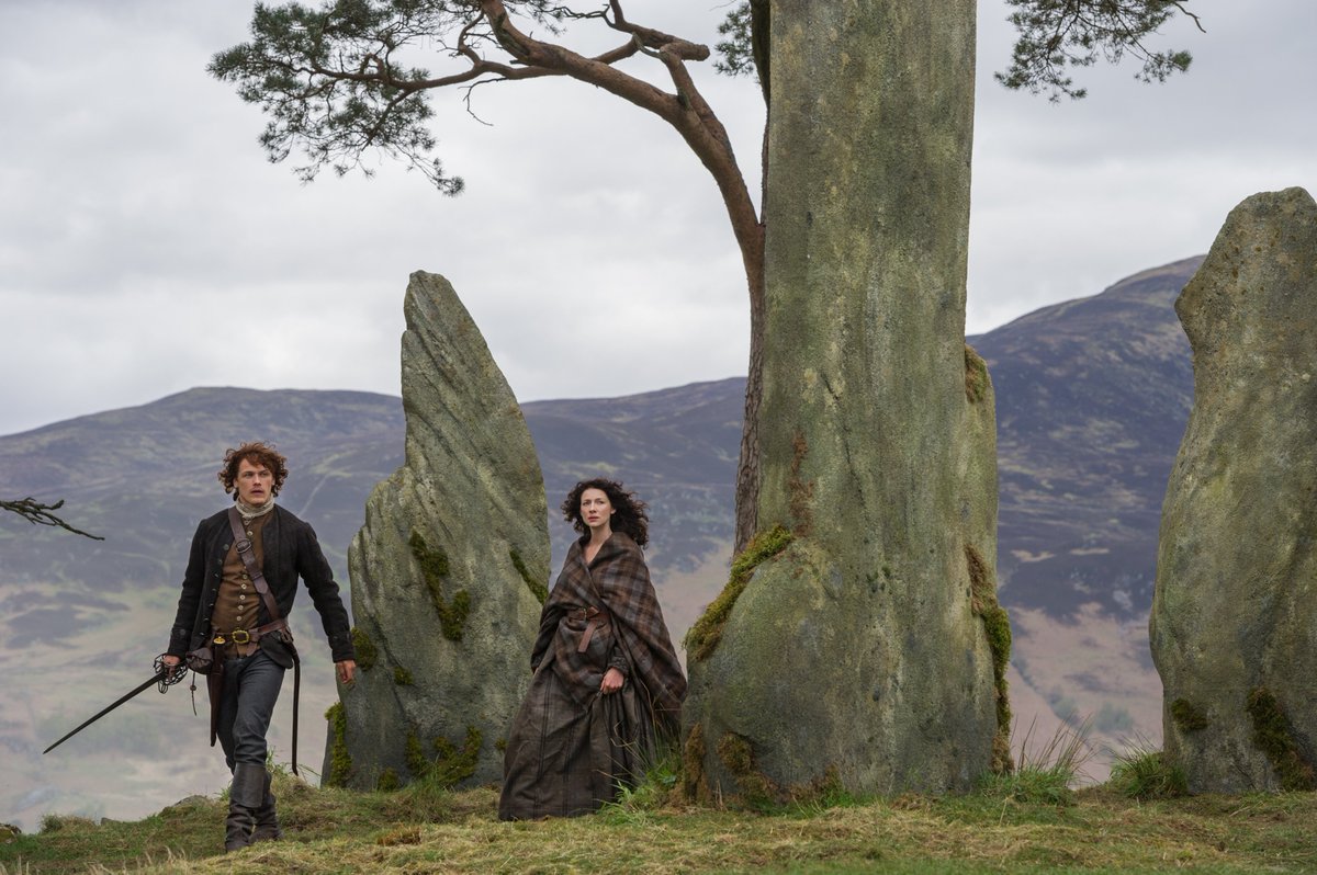 Still from Outlander with the lead characters standing next to a tree and large stones.