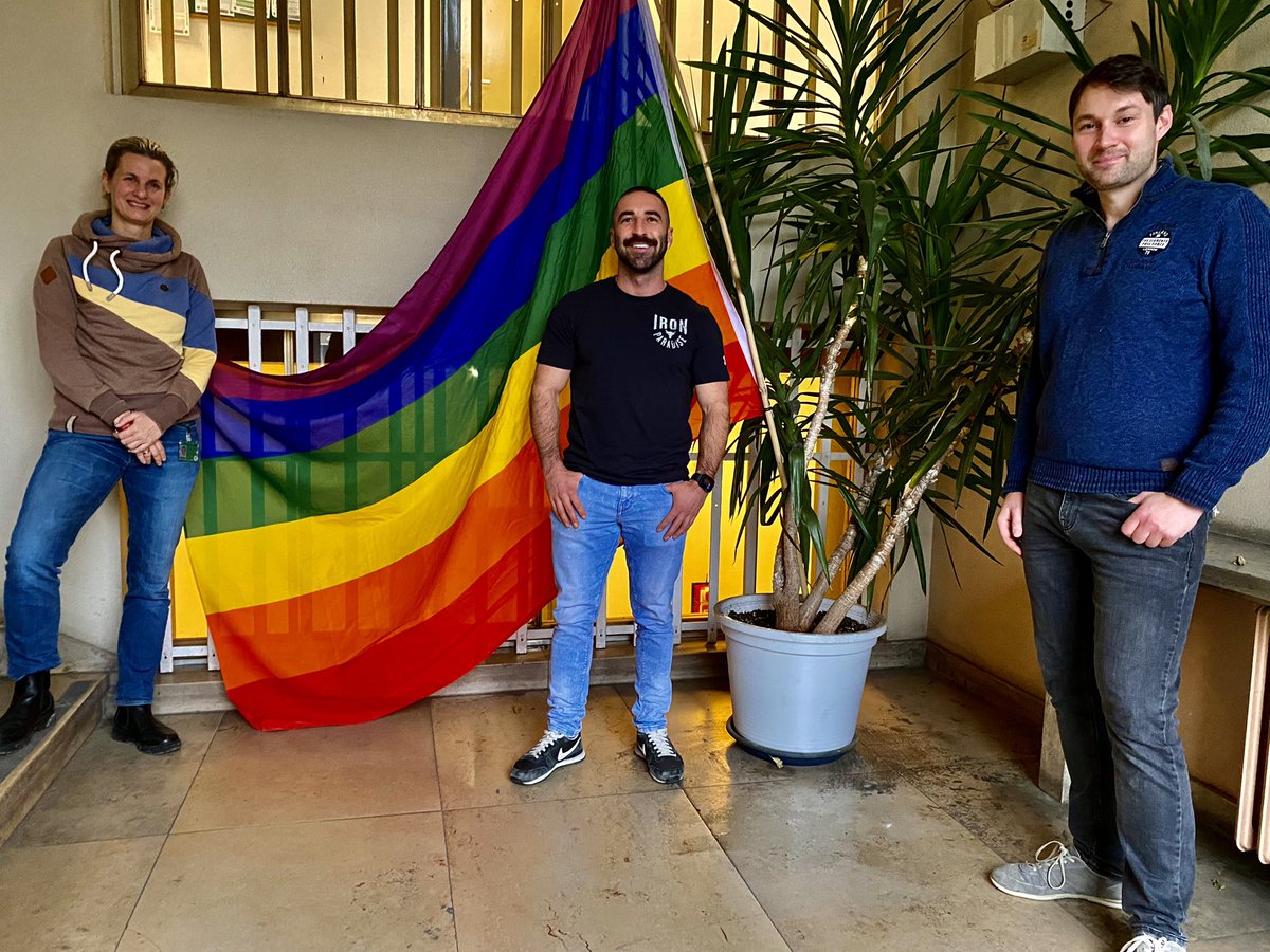 Drei Mitarbeitende der Polizei Berlin in ziviler Kleidung in einem Foyer. Im Hintergrund ist eine Regenbogenfahne zu sehen.