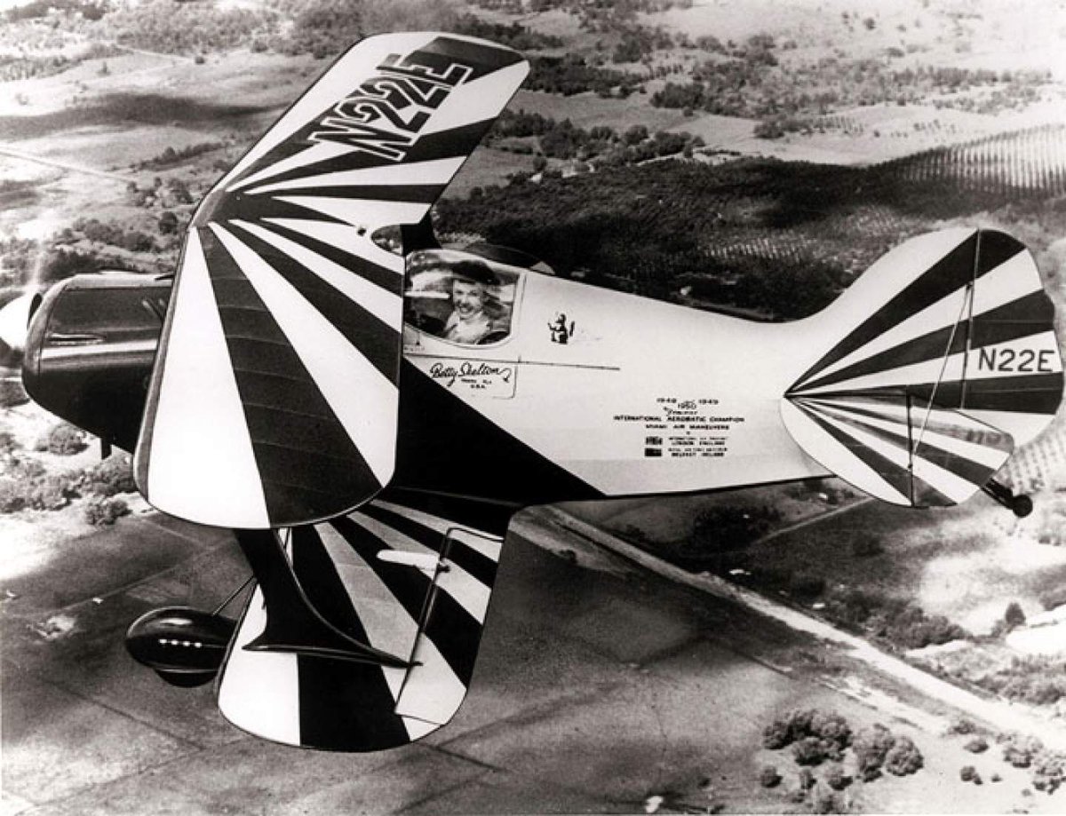 Black and white image of Betty Skelton in the cockpit of an aircraft with black stripes, flying above visible landmarks.