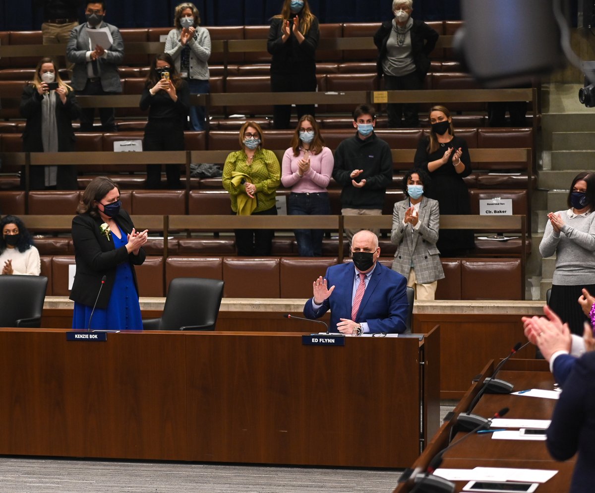 Ed Flynn sits at his desk and waves as members of the City Council and people sitting in the audience seats stand and clap.