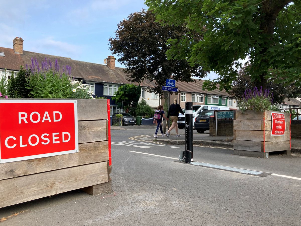 A photo of a wooden road block, holding plants in it, with a red “road closed” sign. In the background of the photo, there’s a child and an adult walking down a street, different coloured houses, and trees.