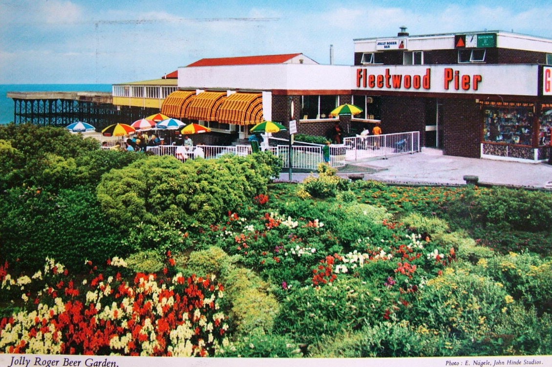 SeasideFerry's tweet image. The Jolly Roger Beer Garden at #Fleetwood #Pier as captured in a #JohnHinde postcard. The garden balances the colours of the bright parasols and #1970s brown/orange awnings shading the beer drinkers. Another satisfying composition @JohnHindeImages @PiersSociety