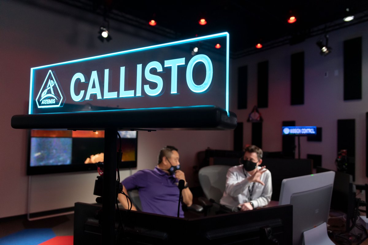 A sign that reads "Callisto" in glowing blue letters in seen in the foreground. In the background, two people wearing masks and headsets converse. 