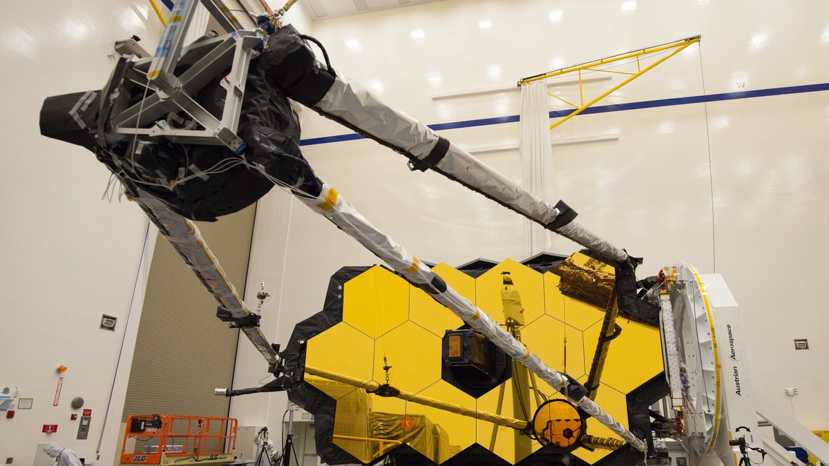 The James Webb Space Telescope shown in a clean room with its secondary mirror, a tripod-like structure, extended out in front of it's unfolded primary mirror. 