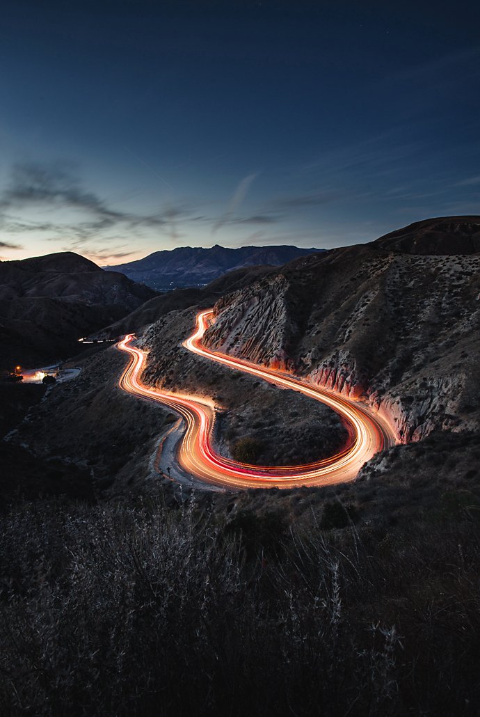 A photograph of a mountain road just as the sunsets. There's a light trail on the road.