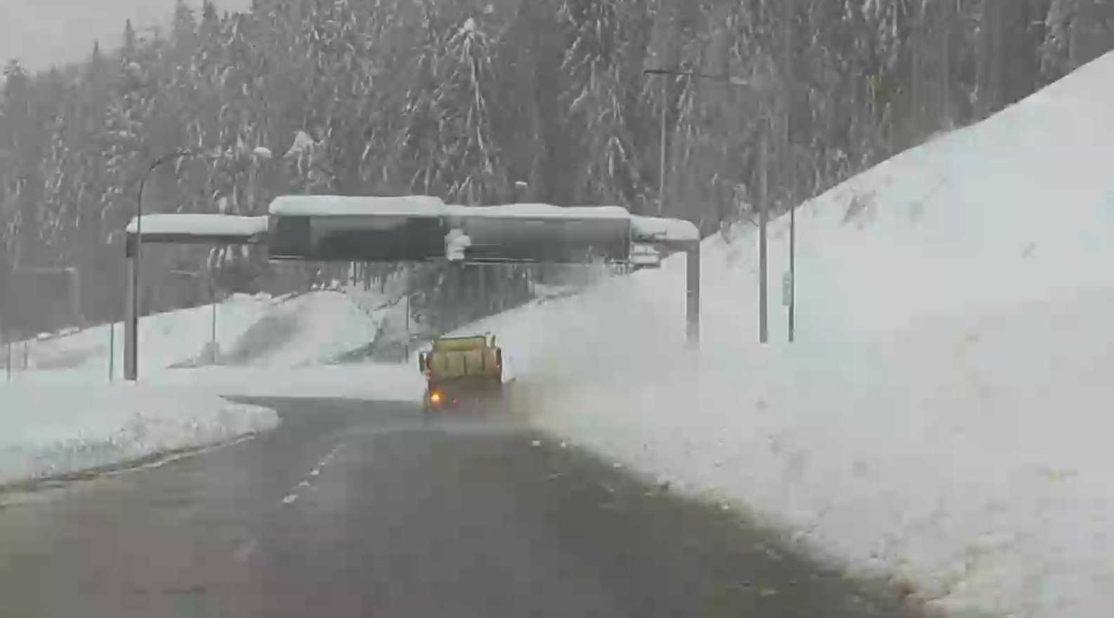 A mountain pass road with two lanes has a large snow plow truck working underneath a large overhead sign that is covered in snow. On each side of the road are huge amounts of snow and there are tons of snow-covered tall trees in the distance.
