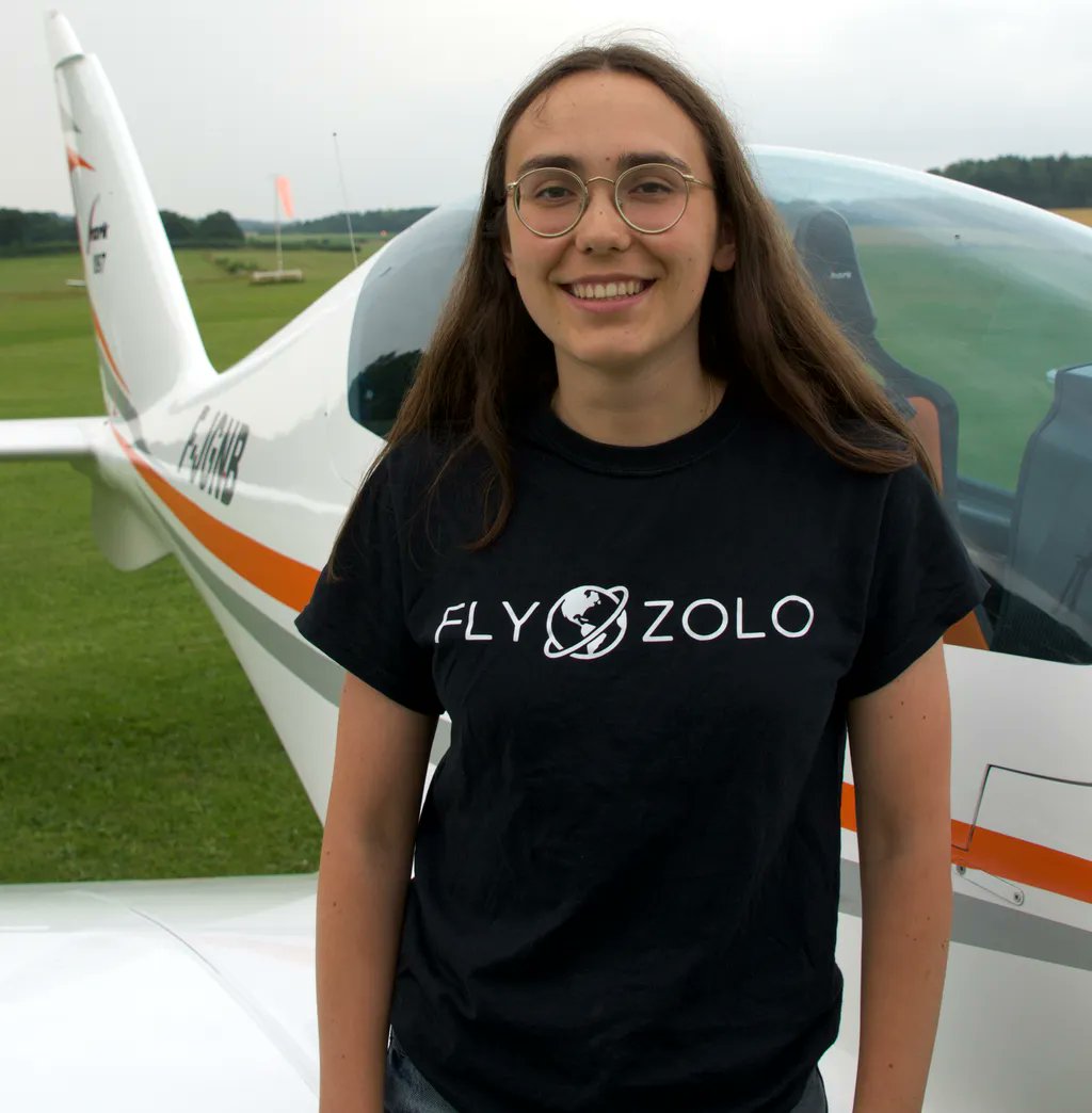 Zara Rutherford, wearing a black t-shirt that says "Fly Zolo" stands in front of a white plane that is on a field of grass