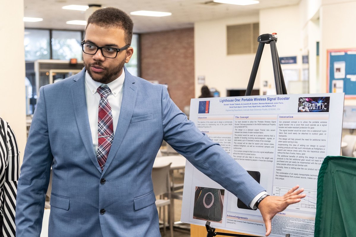 An African American student with glasses and a light blue or grayish suit points to a poster board during a poster session.
