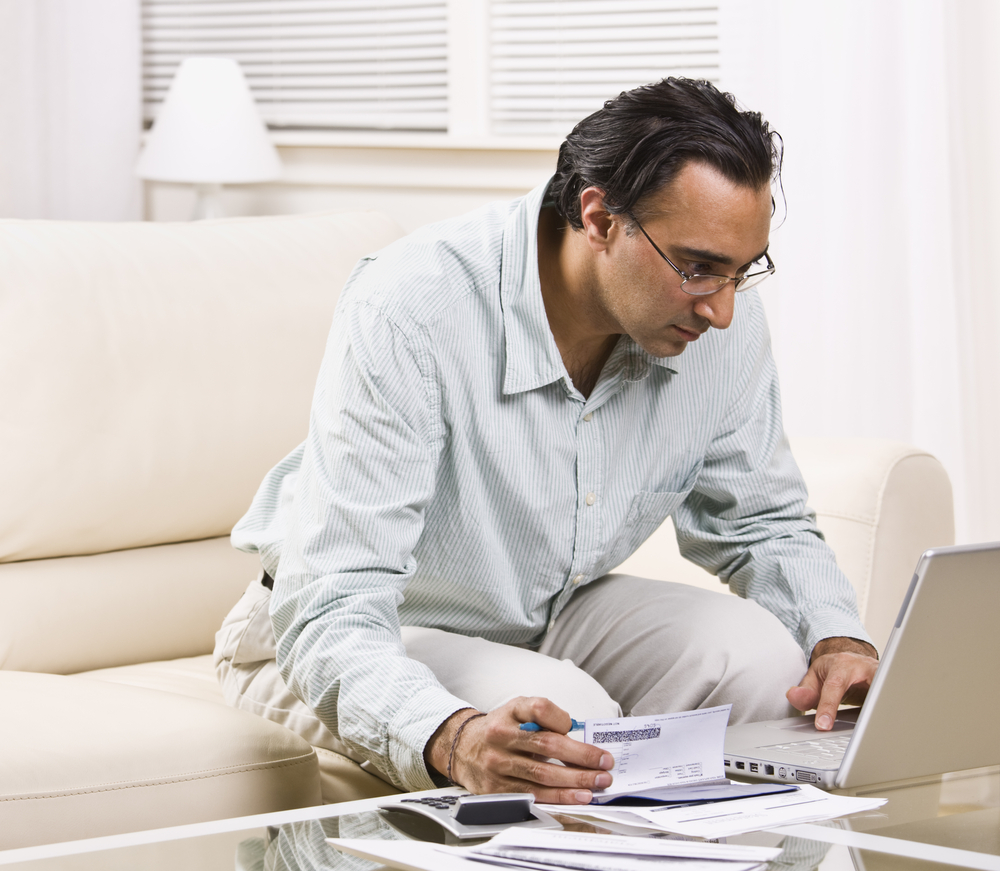 Photo of South Asian man looking at unpaid bills and browsing on his computer .