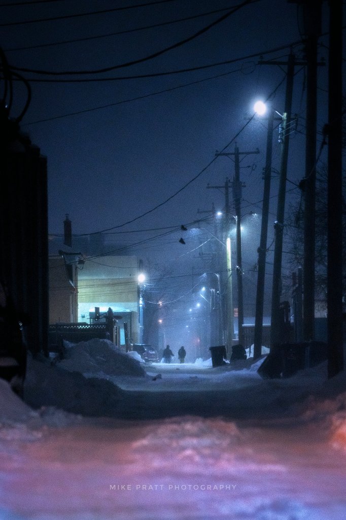 Two figures walk in the middle of a alleyway in an old residential neighborhood. Light snow falls. The mood is very atmospheric.