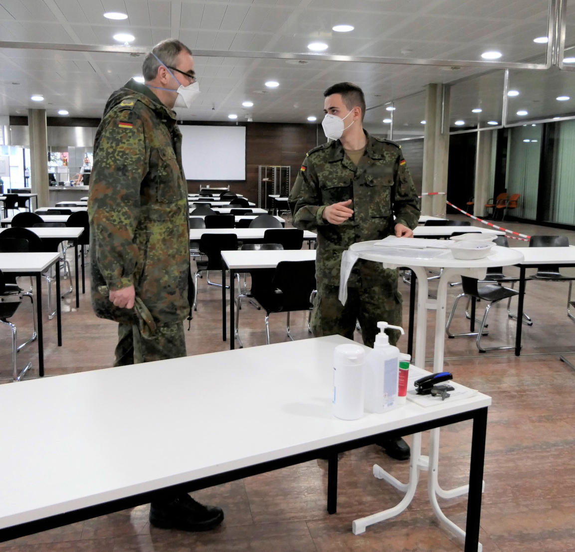 Kommandeur Webert unterhält sich mit einem seiner Soldaten. Beide stehen in einer Mensa, überall sind Tische und Stühle zu sehen.