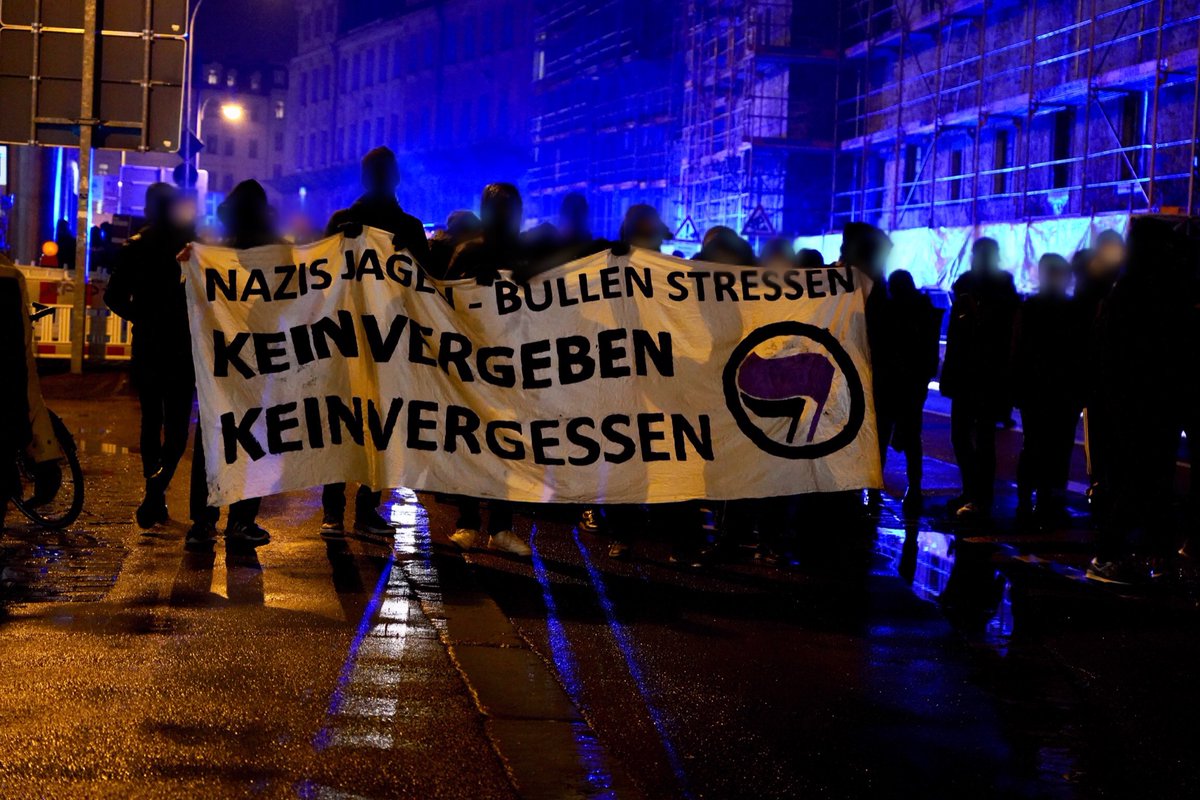 Mehrere Antifas halten ein weißes Transpi mit der Aufschrift „Nazis jagen, Bullen Stressen, kein Vergeben, kein Vergessen“ und einem aufgemalten Antifa Logo (Lila-Schwarz). Hinter den Antifas leuchtet Blaulicht