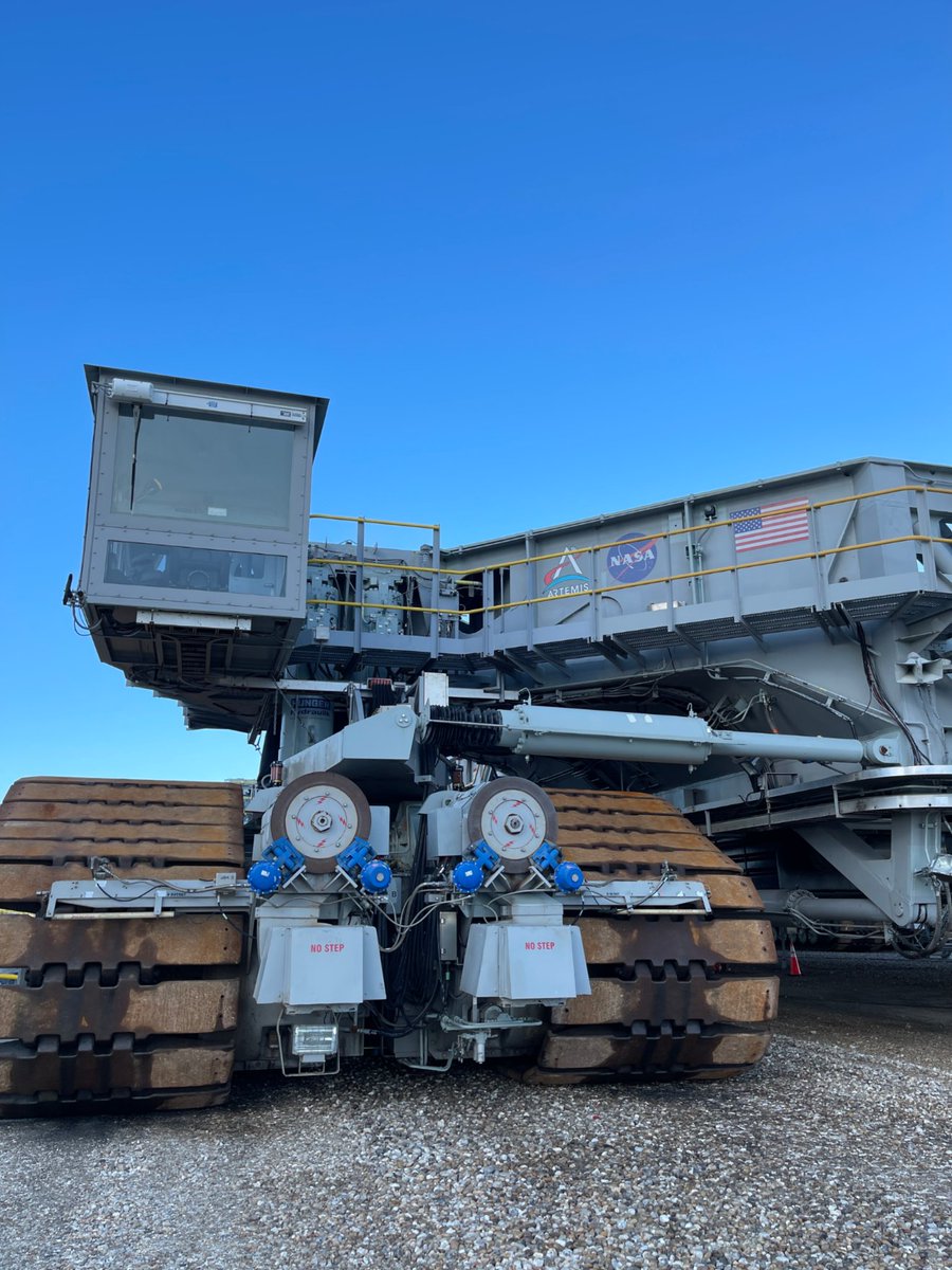 A view of Crawler-Transporter 2 from several meters away. An Artemis logo, NASA insignia, and American flag are visible on the side of the vehicle. 