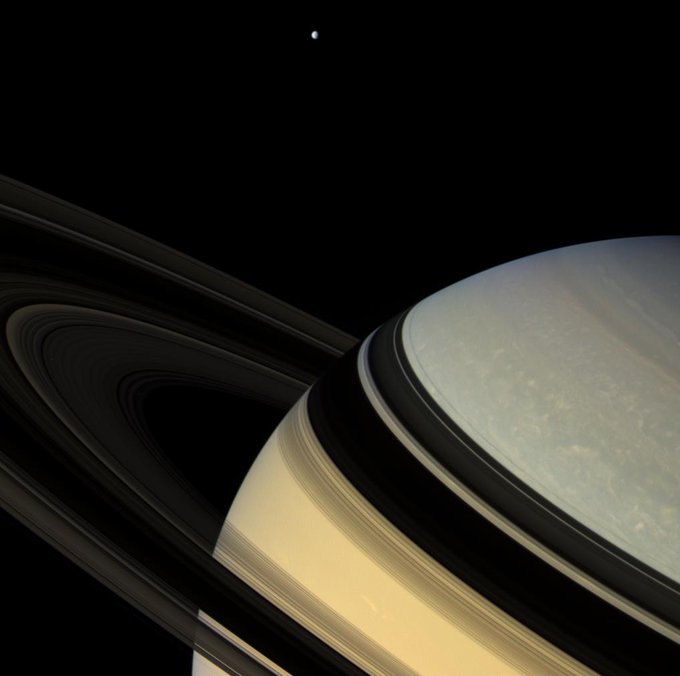 Saturn's rings cast dark banded shadows on the planet's cloud tops. A relatively tiny moon can be seen floating nearby.