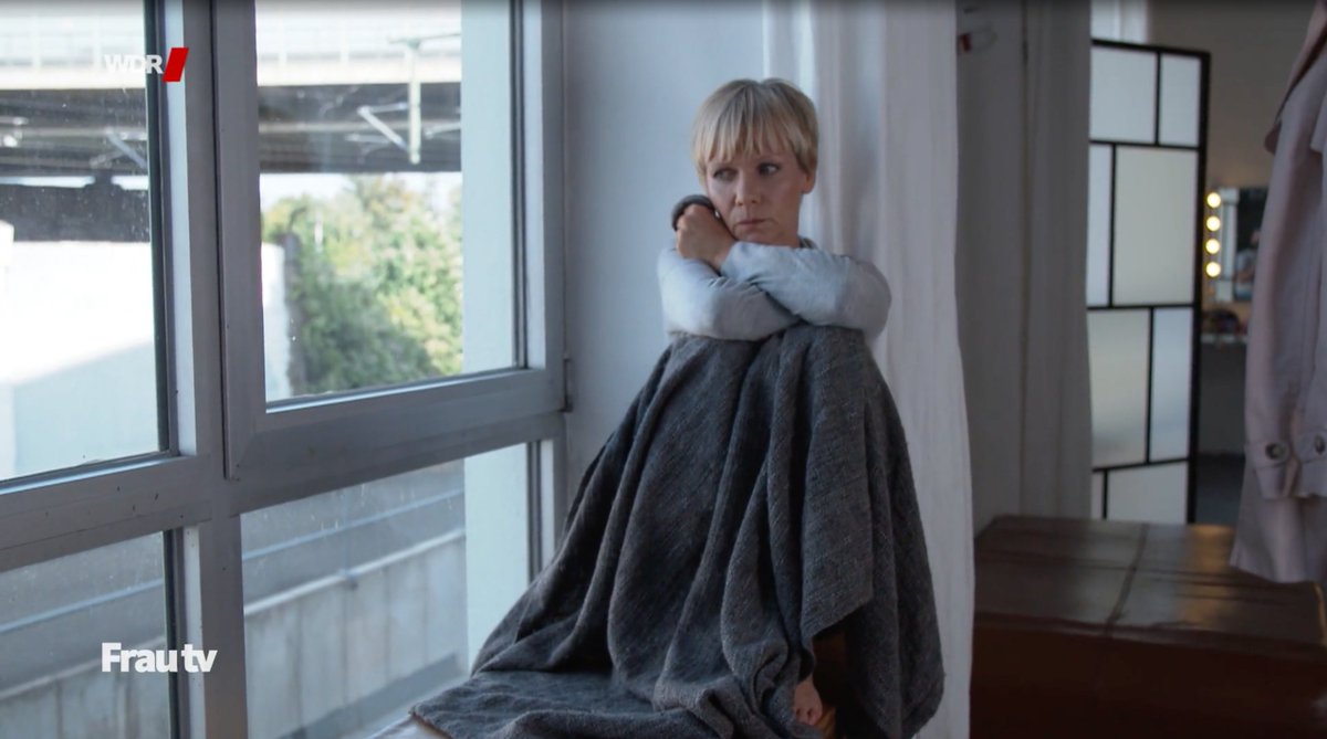 Standbild aus dem Beitrag: Eine Frau sitzt mit besorgtem Blick in eine Decke gehüllt an einem Fenster. 