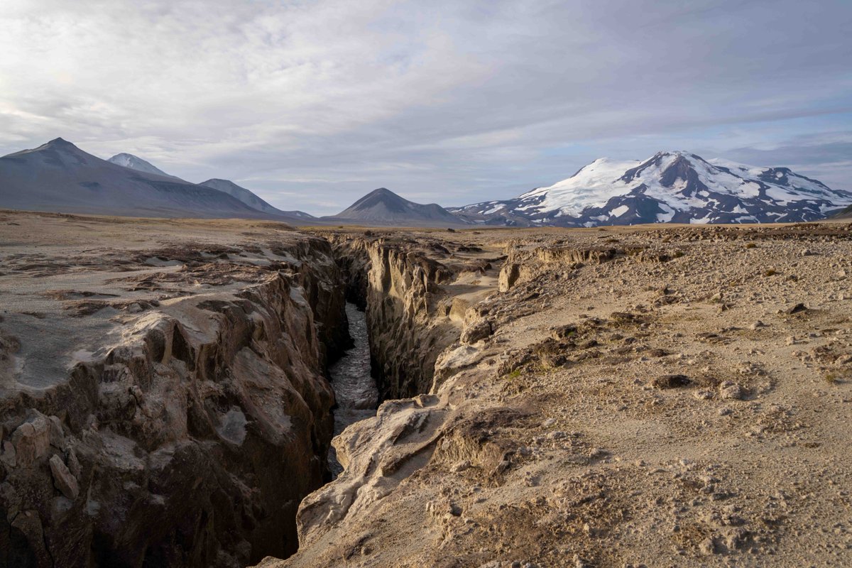 An ashen landscape with a river cutting cutting through and mountains in the background, one mountain is snow and glacier covered