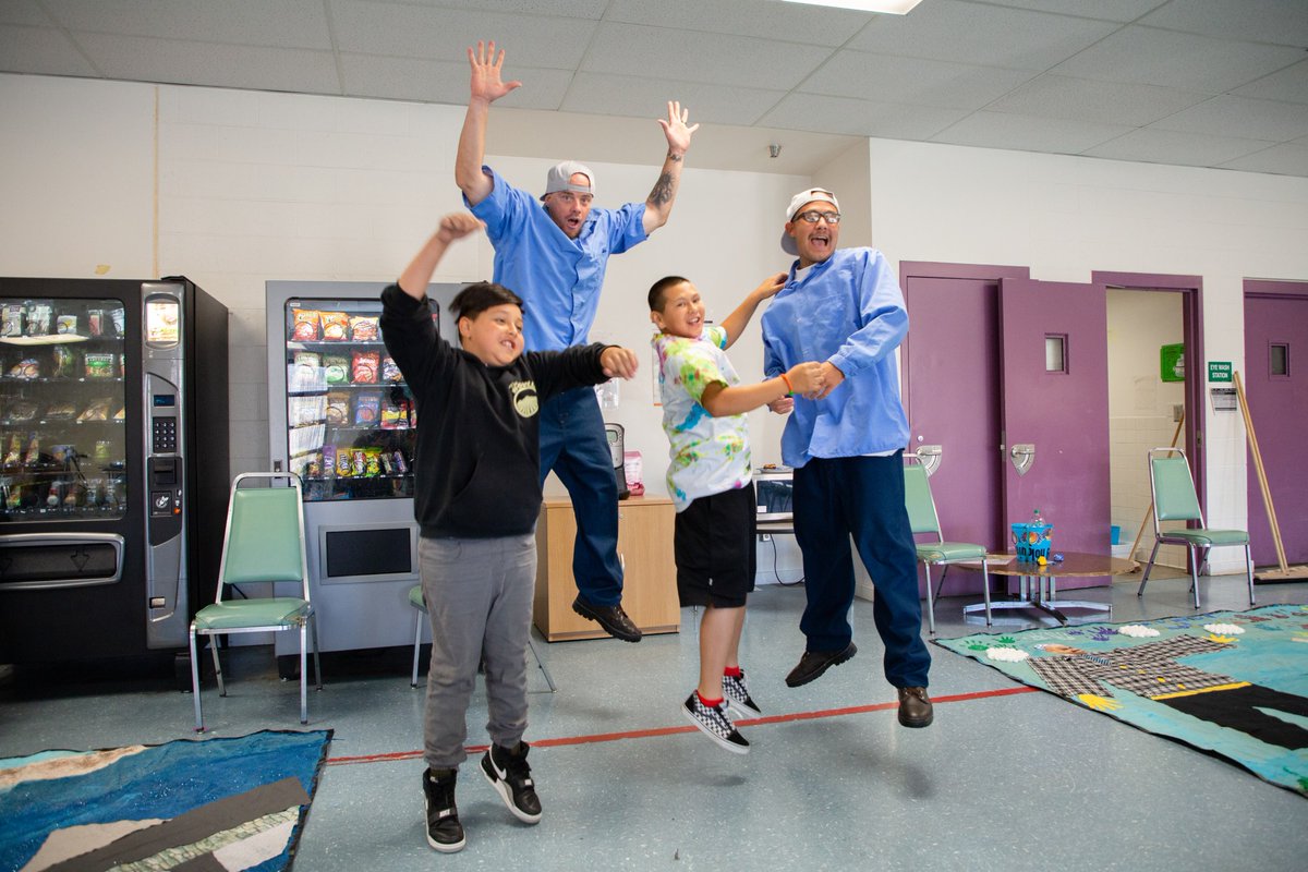 Two fathers, wearing uniform blue shirts and baseball caps, jump excitedly, mid-cheer, with their two sons or relatives in a recreation room.