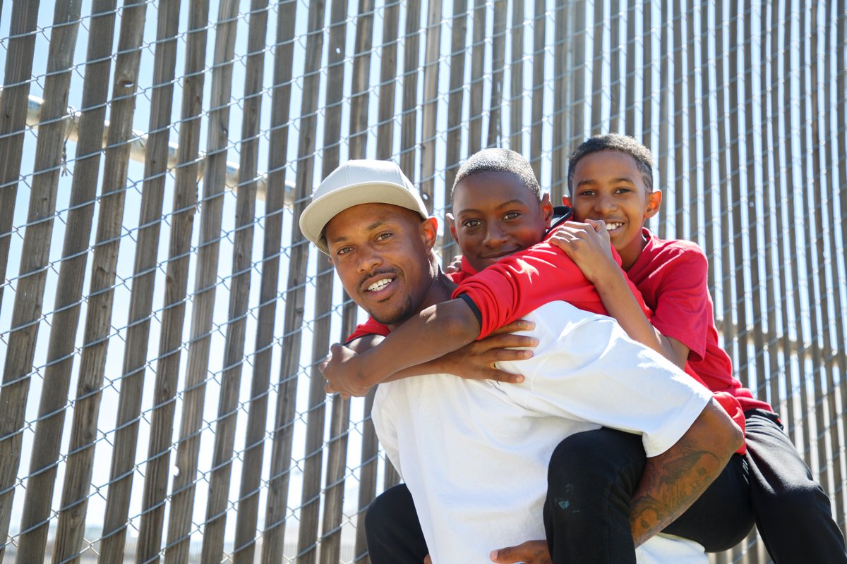 A Black man in a white tee shirt and baseball cap gives a piggy-back ride to his two adolescent sons on a sunny day near a metallic fence. All are smiling.