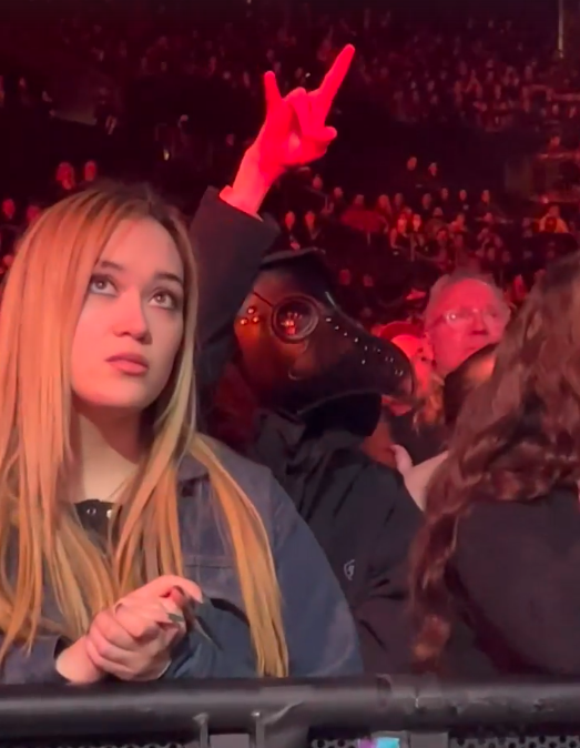 Someone in a plague doctor mask raising the horns sign while the girl in front clasps her hands together and has a facial expression that makes it look like she's praying.