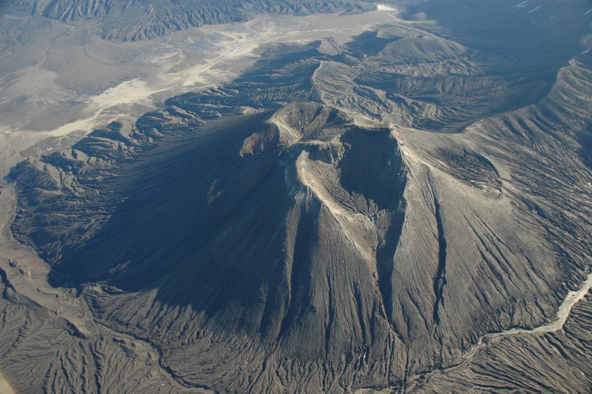 A black volcanic cone with two craters at the top, with deep erosional valleys down the flanks.
