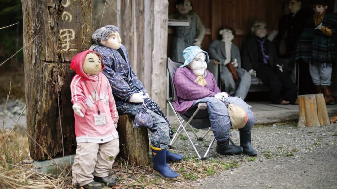 A photo of some scarecrows, dressed as various types of person, sitting around outside what looks like a tea house or shop.