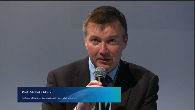 Michel is in a suit and has a microphone in his hand as he presents. in the bottom left, there is a box that says his name and 'professor of fisheries conservation at Heriot Watt Univeristy'.