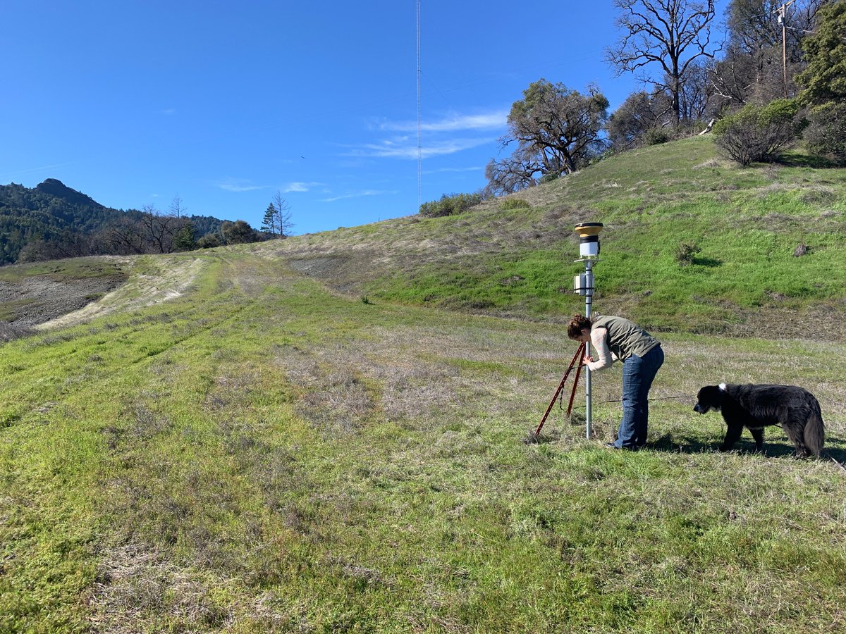 A women scientist checks on a research instrument in a grassy field. She is accompanied by an adorable black dog.