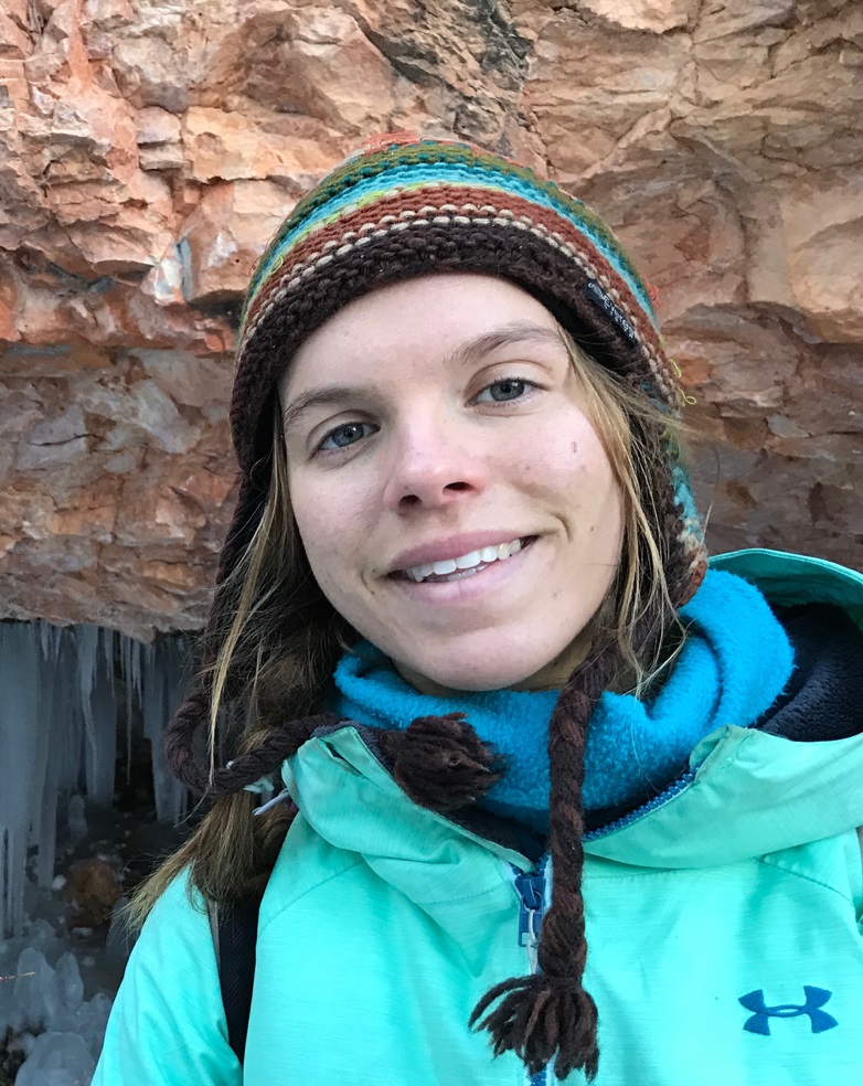 A smiling woman wearing a beanie stands near a rock.