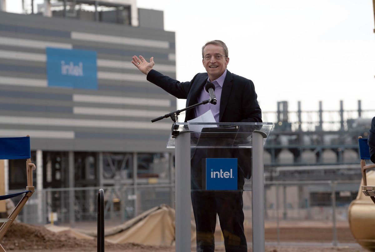 Pat Gelsinger stands at a podium while speaking into a microphone and gesturing at a factory behind him. 