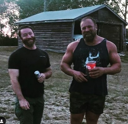 Image of two men smiling and standing in a rural area in front of a shed. Left: Jeremy MacKenzie, holding a canned drink, wearing a black t-shirt and green pants. On the right is a taller, muscular man wearing a black tank top with a white design and camouflage shorts. He is holding a Big Gulp drink.