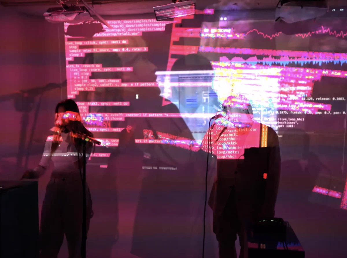 A still from the video Araya, DJ_Dave - Kiss on the Train (Live Coded Video). 

DJ Dave and Araya are standing in front of lots of projected Sonic Pi code holding mics and singing.