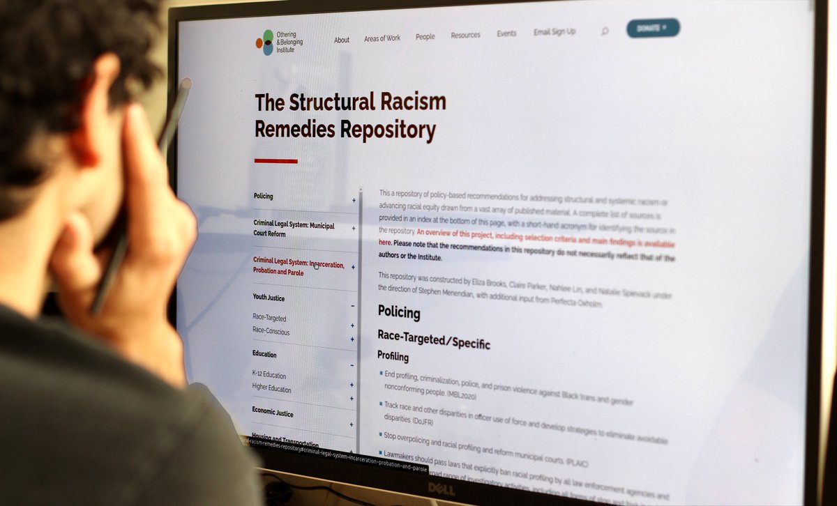Image of a lighter-skin person with dark hair and a sark shirt sitting in front of a computer screen. On the screen is Othering and Belonging Institute at UC Berkeley's "The Structural Racism Remedies Repository".