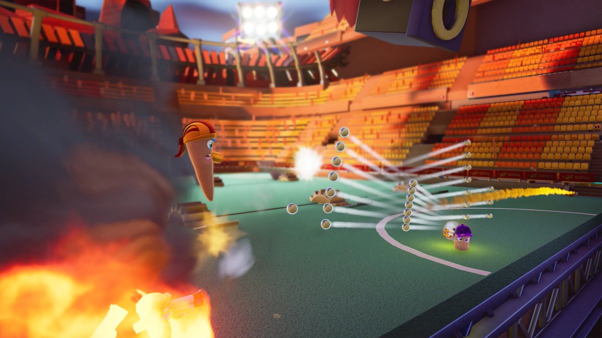 In-game screenshot from Worms Rumble. Shows two Worms firing bullets at each other in the middle of a sports stadium. There are big flames in the corner of the image.