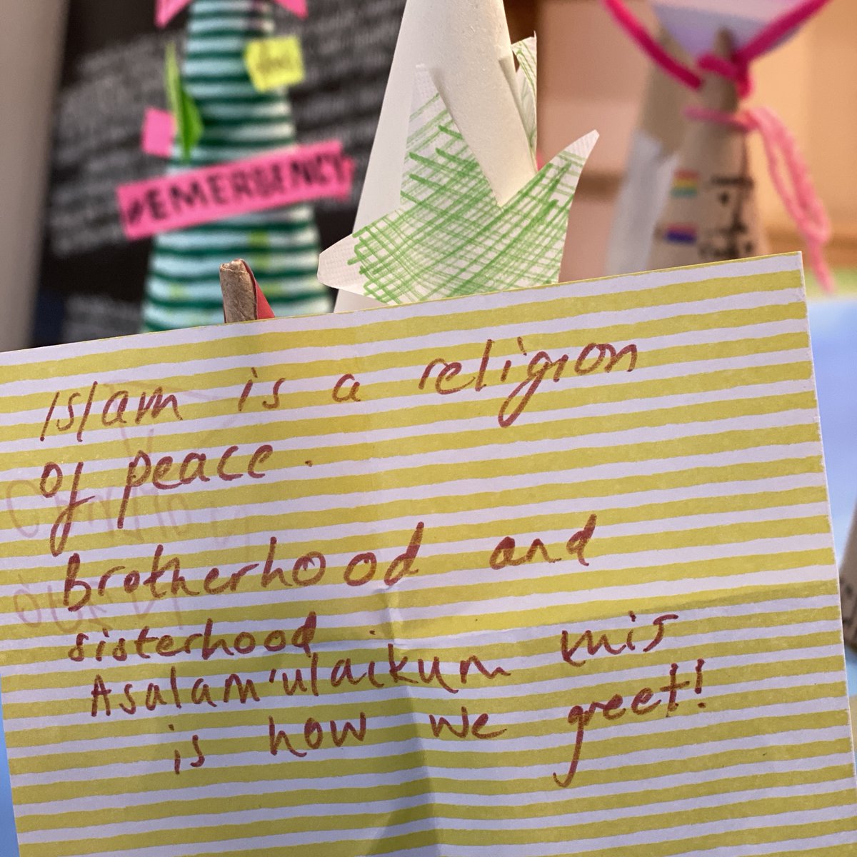 A yellow and white finely striped paper is unfolded and the words, "Islam is a religion of peace. Brotherhood and sisterhood. Asalam'ulaikum this is how we greet!"are written inside. In the background are other artworks that visitors have made but they're blurred and difficult to make out.