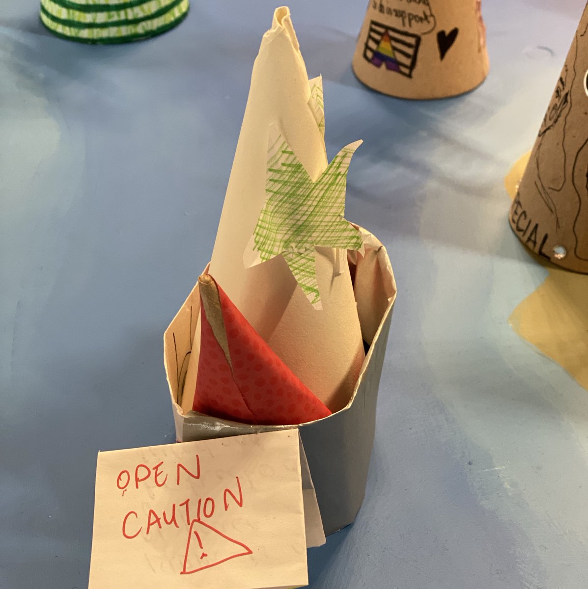 Photo of a small paper boat/mosque. The paper boat has two visible cones within it, the tallest of which has a large star. We can see a folded piece of paper stuck on the boat which has the words, "OPEN CAUTION" The next picture has the message that's contained within the folded paper.