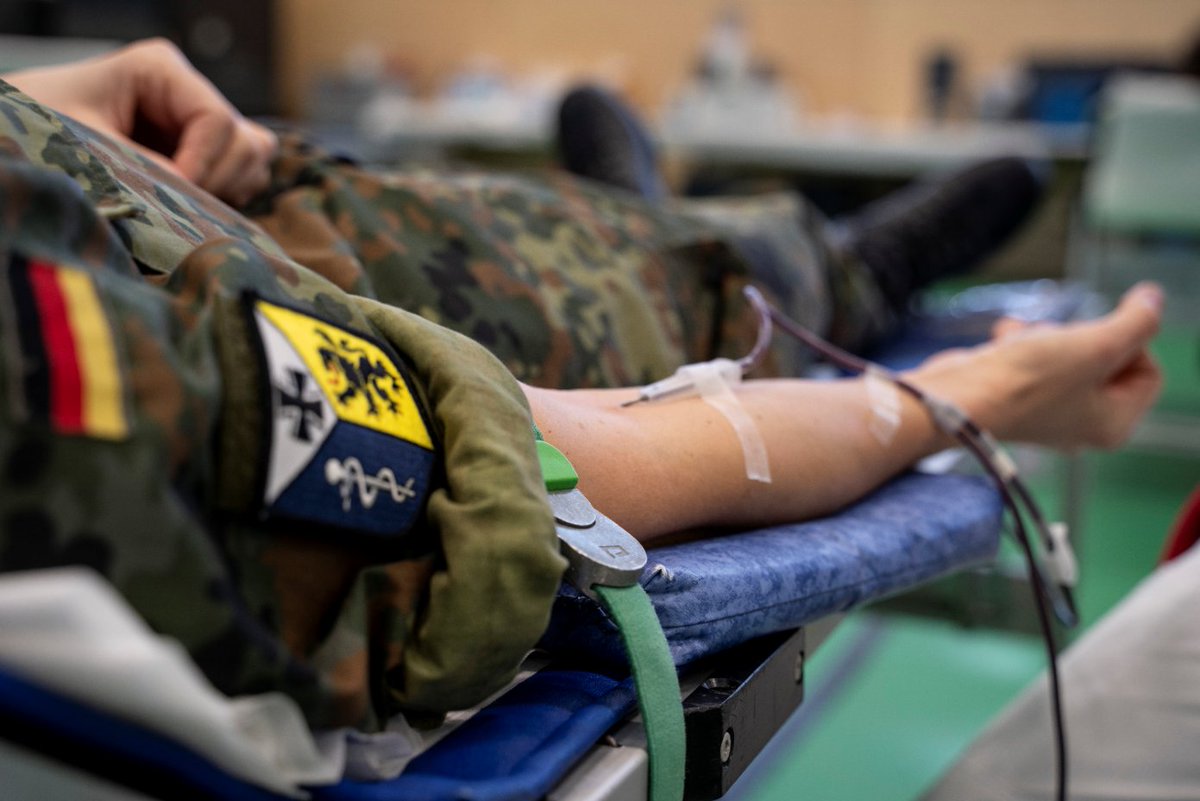 Nahaufnahme eines Armes, in dessen Vene eine Nadel zum Blutspenden steckt. Der Soldat, dem der Arm gehört, trägt eine grüne Uniform, Deutschlandflagge und Wappen des Sanitätsregimentes 3 sind darauf zu sehen.