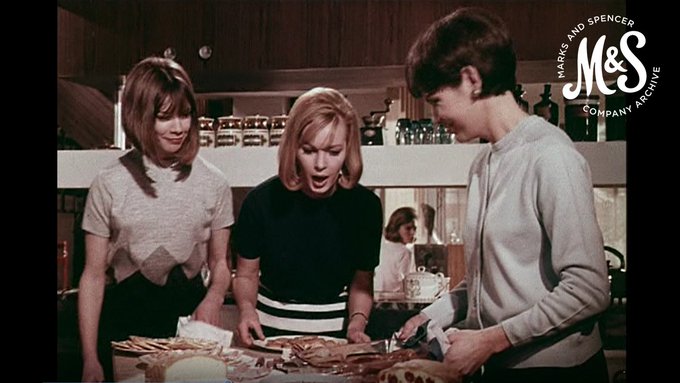 A colour screenshot from a 1960s advert for M&S clothing. Three women stand in an ultra-modern 60s kitchen looking at plates of canapes. 