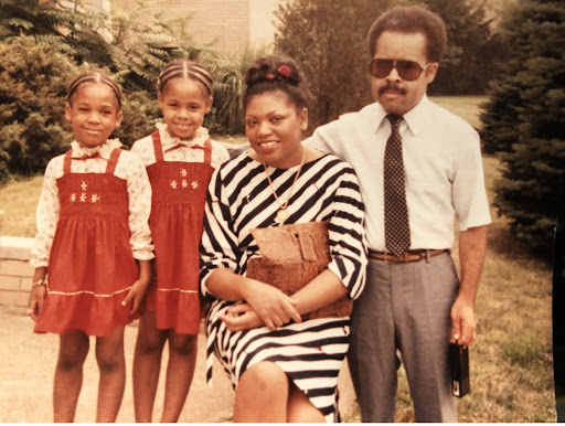 a young, Amoretta Morris, with her sister and parents. the children are wearing red dresses, mom is wearing a black and white stripped dress, and dad is wearing gray slacks, a white button down shirt, and a tie.