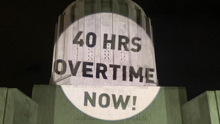 A projection on the capital building in Salem, OR that reads “40 hours overtime now!”