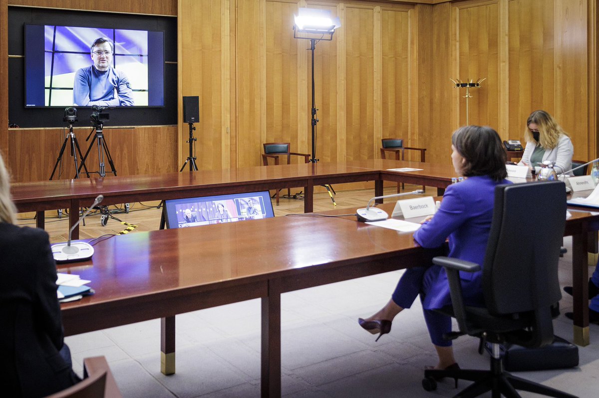 Annalena Baerbock schaut auf einen Monitor, auf dem Dmytro Kuleba zu sehen ist. Sie sitzt an einem langen Holztisch, neben ihr zwei Mitarbeiterinnen, im Auswärtigen Amt.