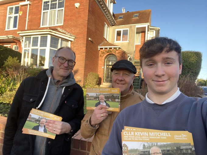 Three people men holding calling cards saying ‘Cllr Kevin Mitchel’ in front of a red brick house in Exeter.