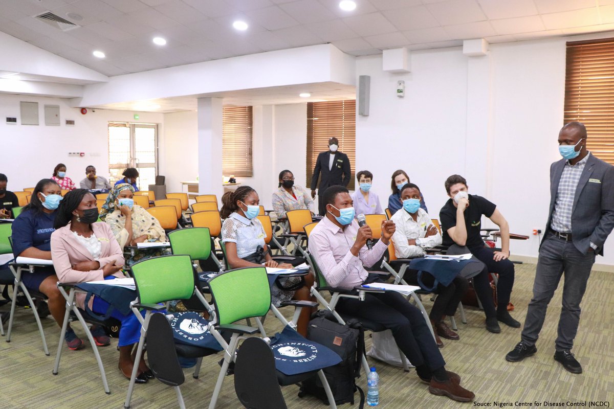 A man in a suit stands in front of a half-filled seminar room and listens to a man with microphone sitting in the front row.