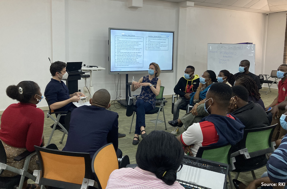 A group of 15 people, who all wear face masks, sits in a seminar room and discusses in front of a large-format screen and a flip chart. (Source: RKI)