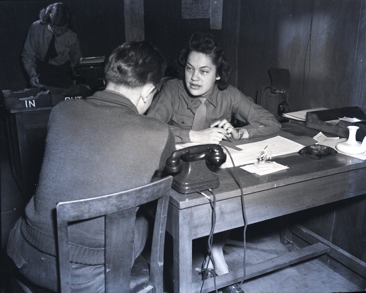 Black and white photograph of a woman in a desk having a conversation with someone.