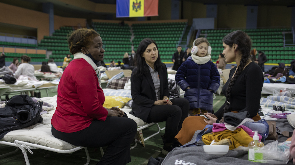 Three women sit talking on camp beds inside an indoor sports arena. A child stands in the middle. The Moldovan flag hangs from the roof of the arena and we can see the rows of seats at the edge of the arena.
